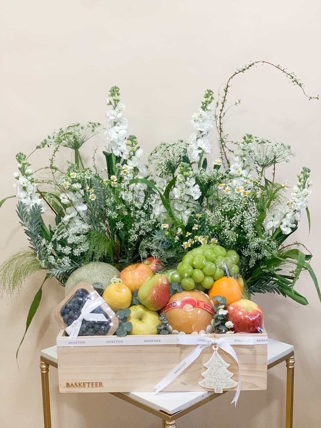 A wooden crate filled with an assortment of fruits including grapes, pears, oranges, a melon, and blueberries. It is decorated with tall white flowers and greenery, and wrapped with a white ribbon labeled 