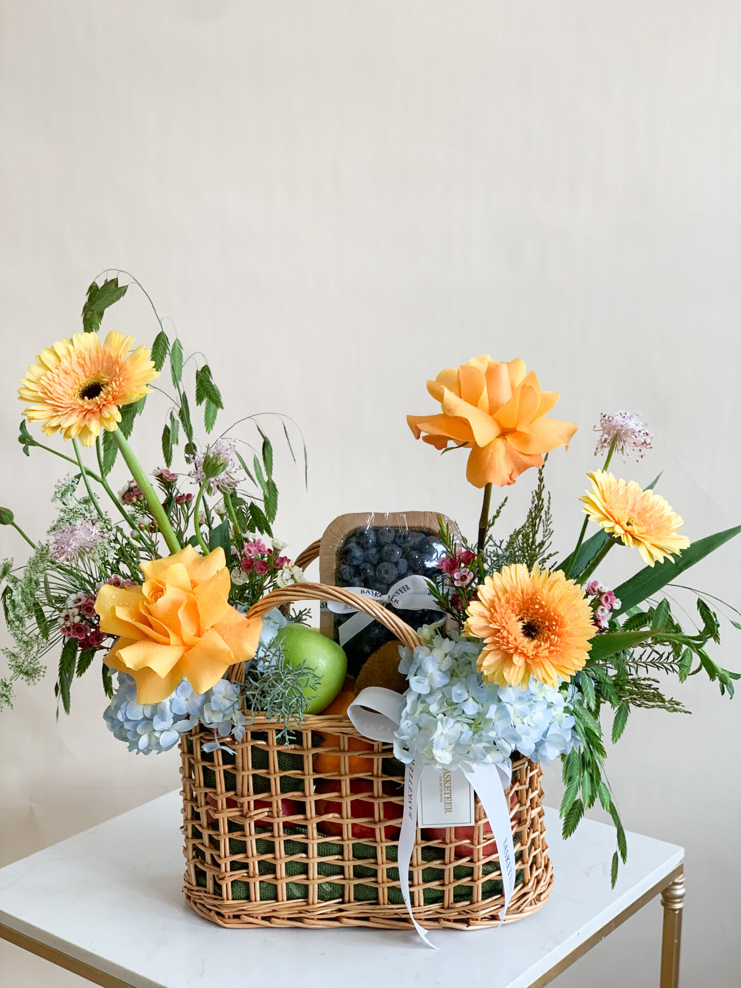 Fresh fruit and flower basket featuring yellow roses, gerberas, blue hydrangeas, and assorted fruits in a woven basket with a white ribbon.