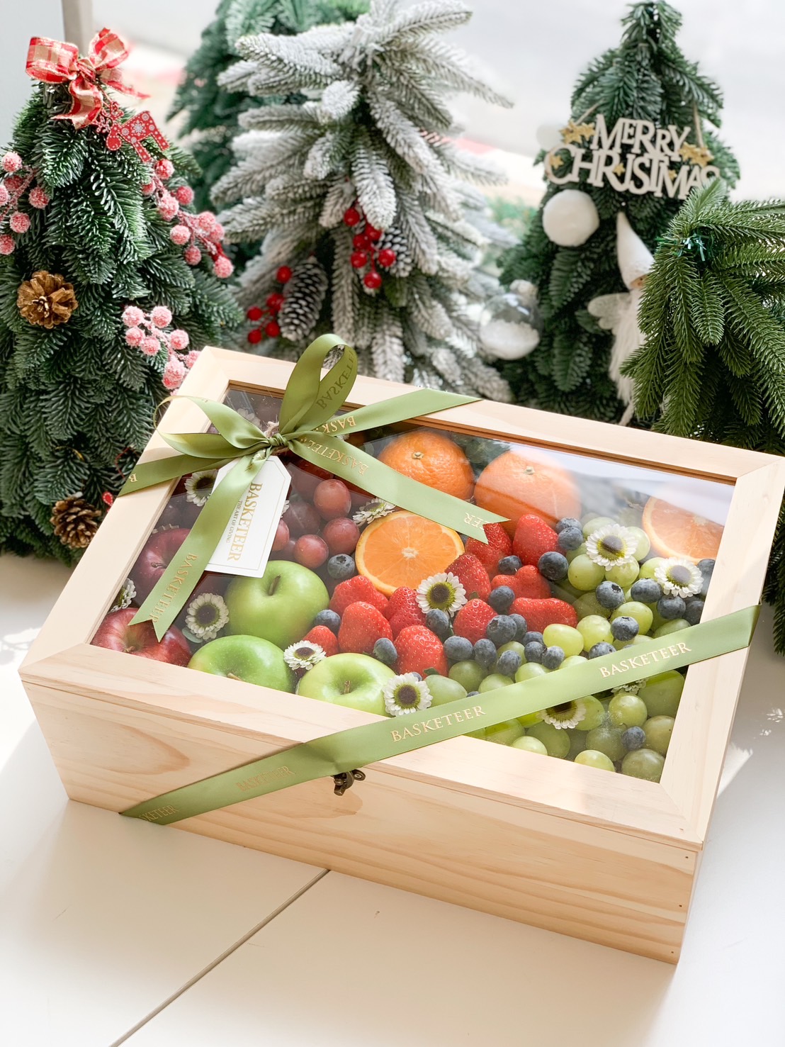 Wooden box of fresh fruits including green apples, grapes, oranges, and berries, adorned with green ribbons, set amidst Christmas decorations.
