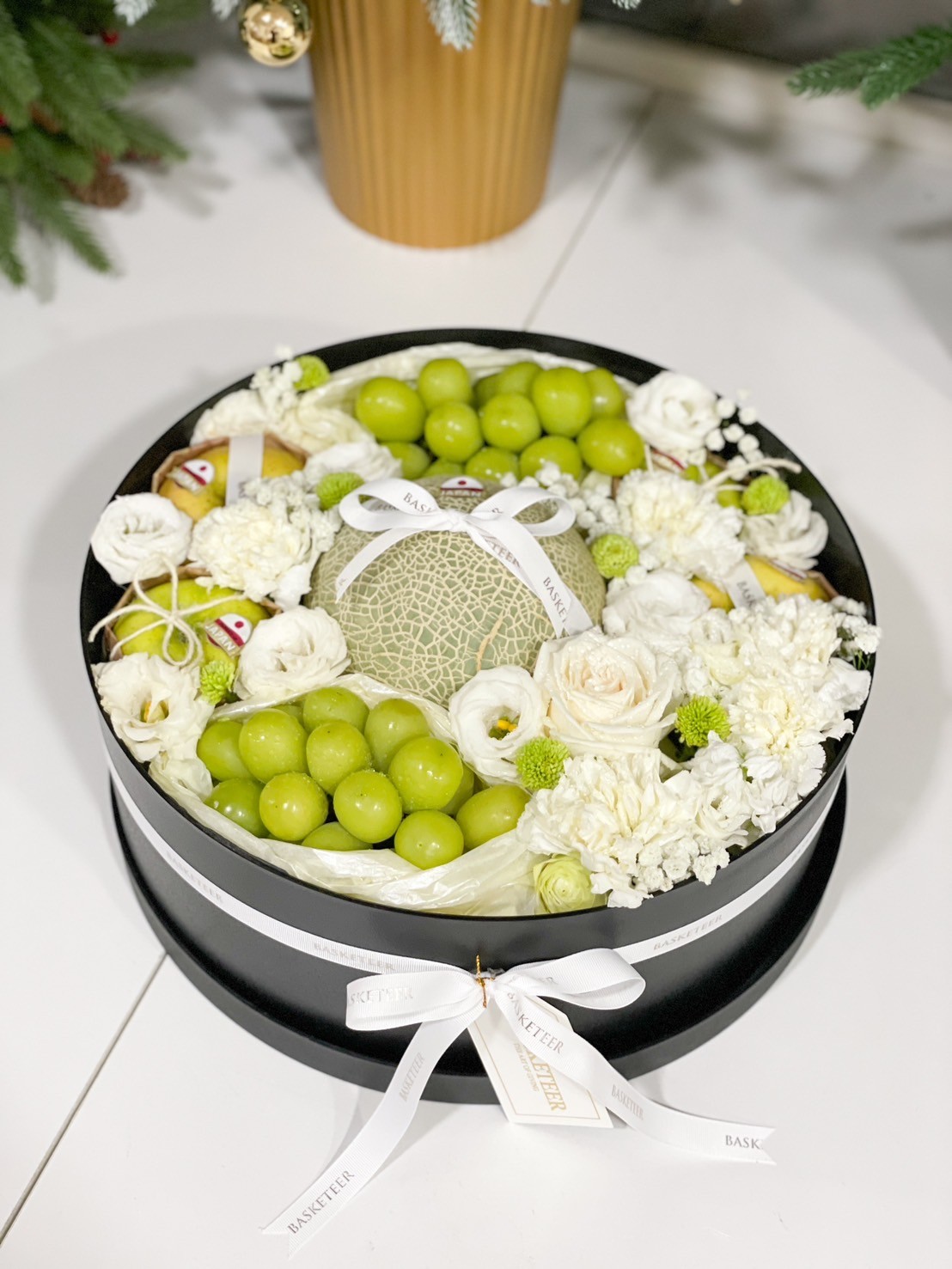Luxury fruit box with premium melon, green grapes, and white floral arrangement, beautifully presented in a black round box.
