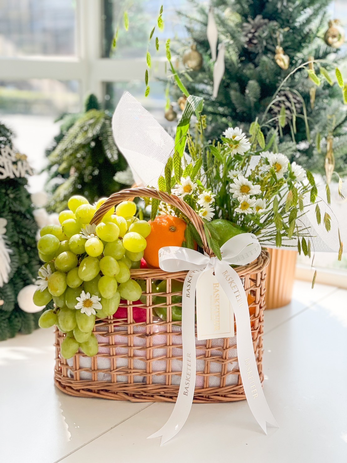Fresh fruit basket with green grapes, oranges, and white daisies, elegantly arranged with a white ribbon for gifting.