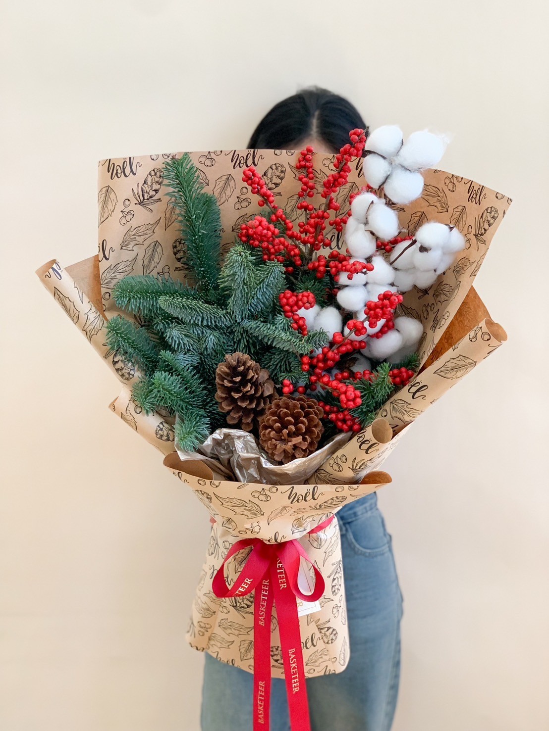 Christmas bouquet with pinecones, red berries, cotton stems, and green pine leaves, wrapped in festive Noel-themed paper.