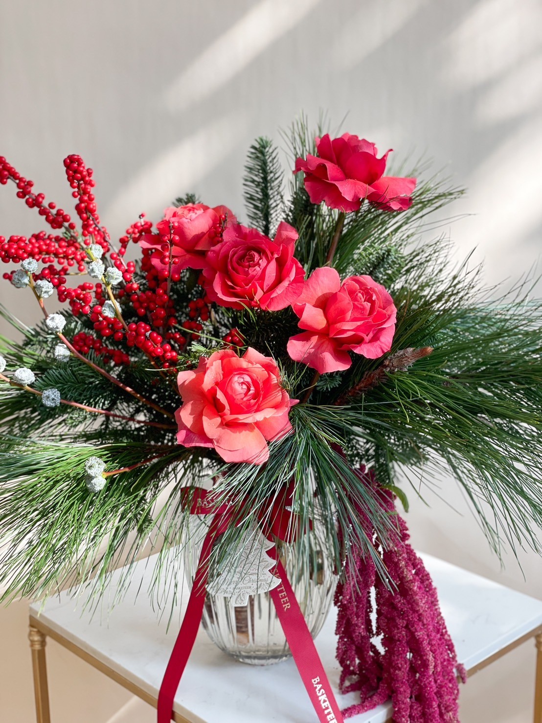 Christmas vase arrangement featuring red roses, pine leaves, red berries, and decorative holiday ribbons in a glass vase.