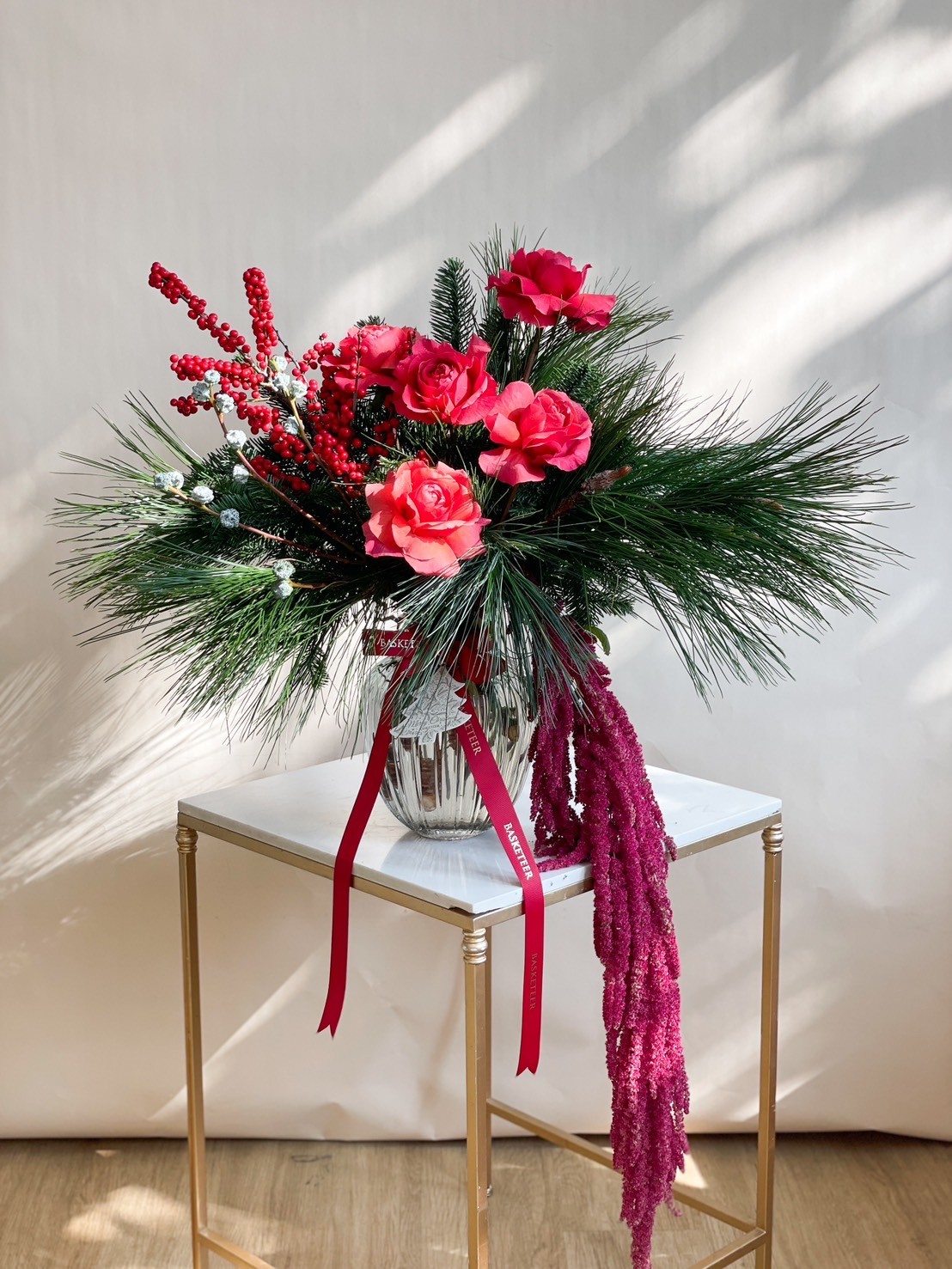 Christmas vase arrangement featuring red roses, pine leaves, red berries, and decorative holiday ribbons in a glass vase.