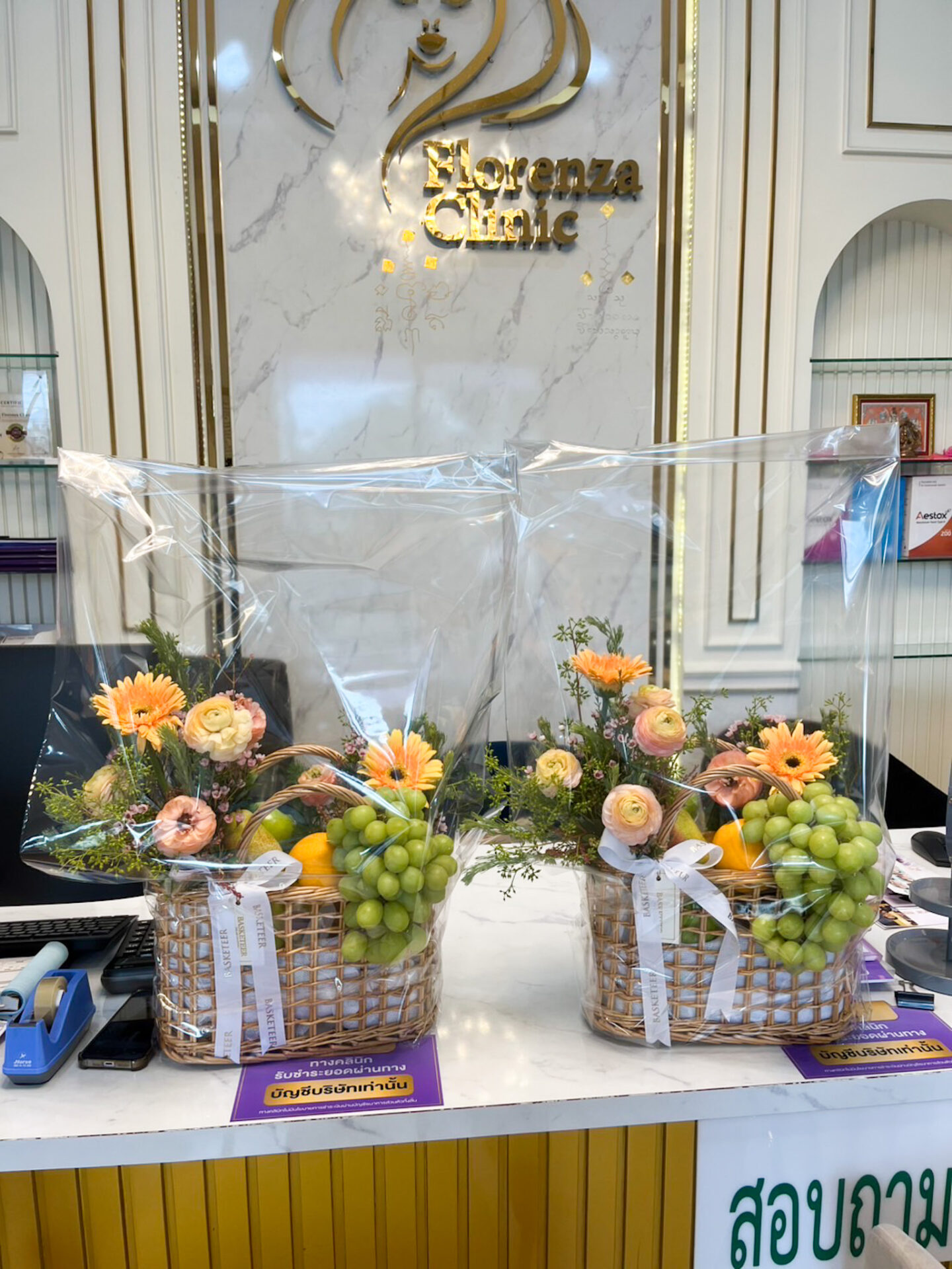 Luxury fruit basket with green grapes, oranges, and assorted fruits, decorated with peach ranunculus, orange gerbera, and seasonal flowers, wrapped with satin ribbon.