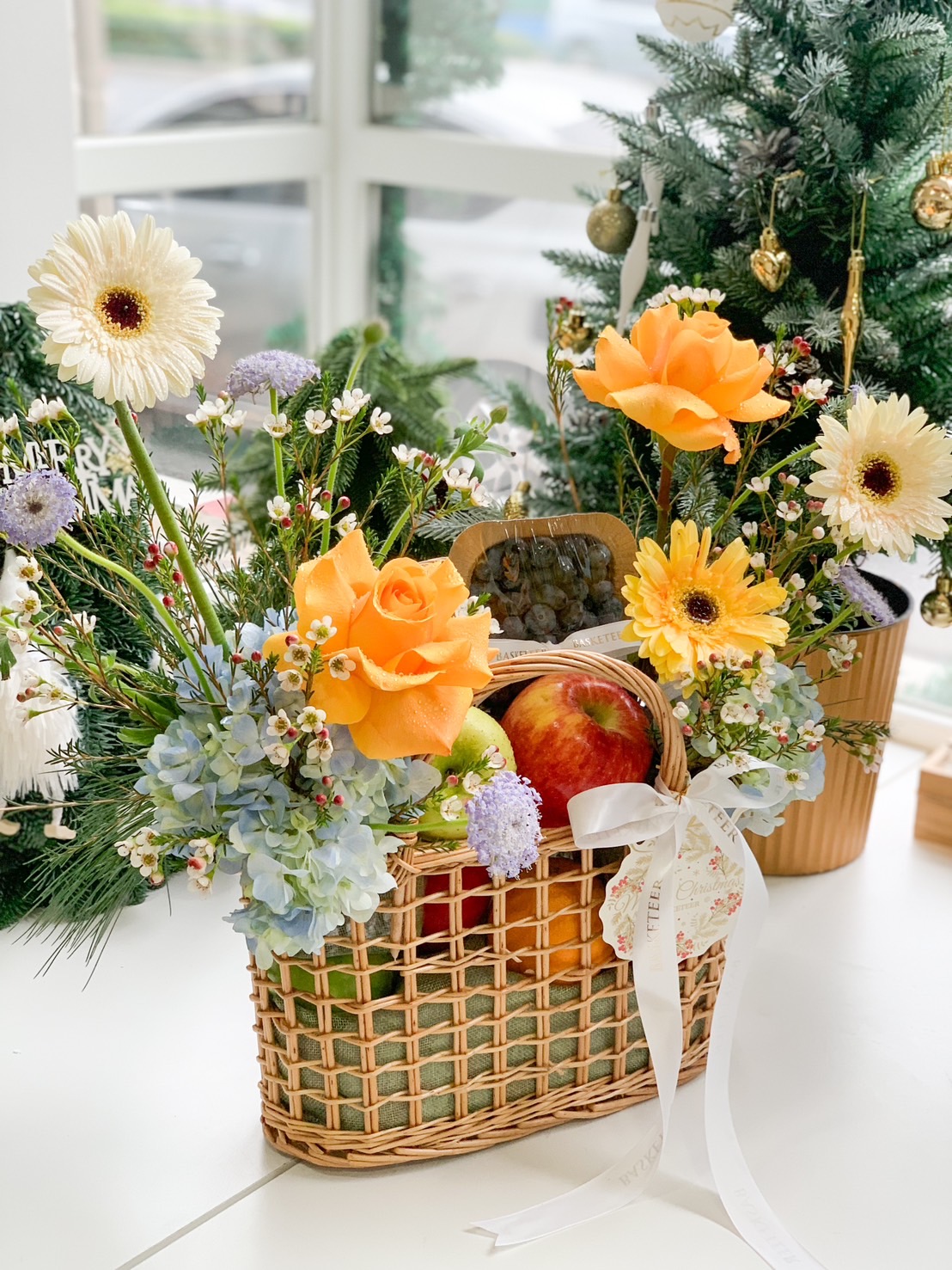 A beautifully crafted wicker basket filled with fresh fruits, including apples, lemons, and kiwis, accented with vibrant floral arrangements featuring orange tulips, blue hydrangeas, and yellow sunflowers, placed on a white table with scattered fruit decorations, perfect for gifting or a centerpiece.