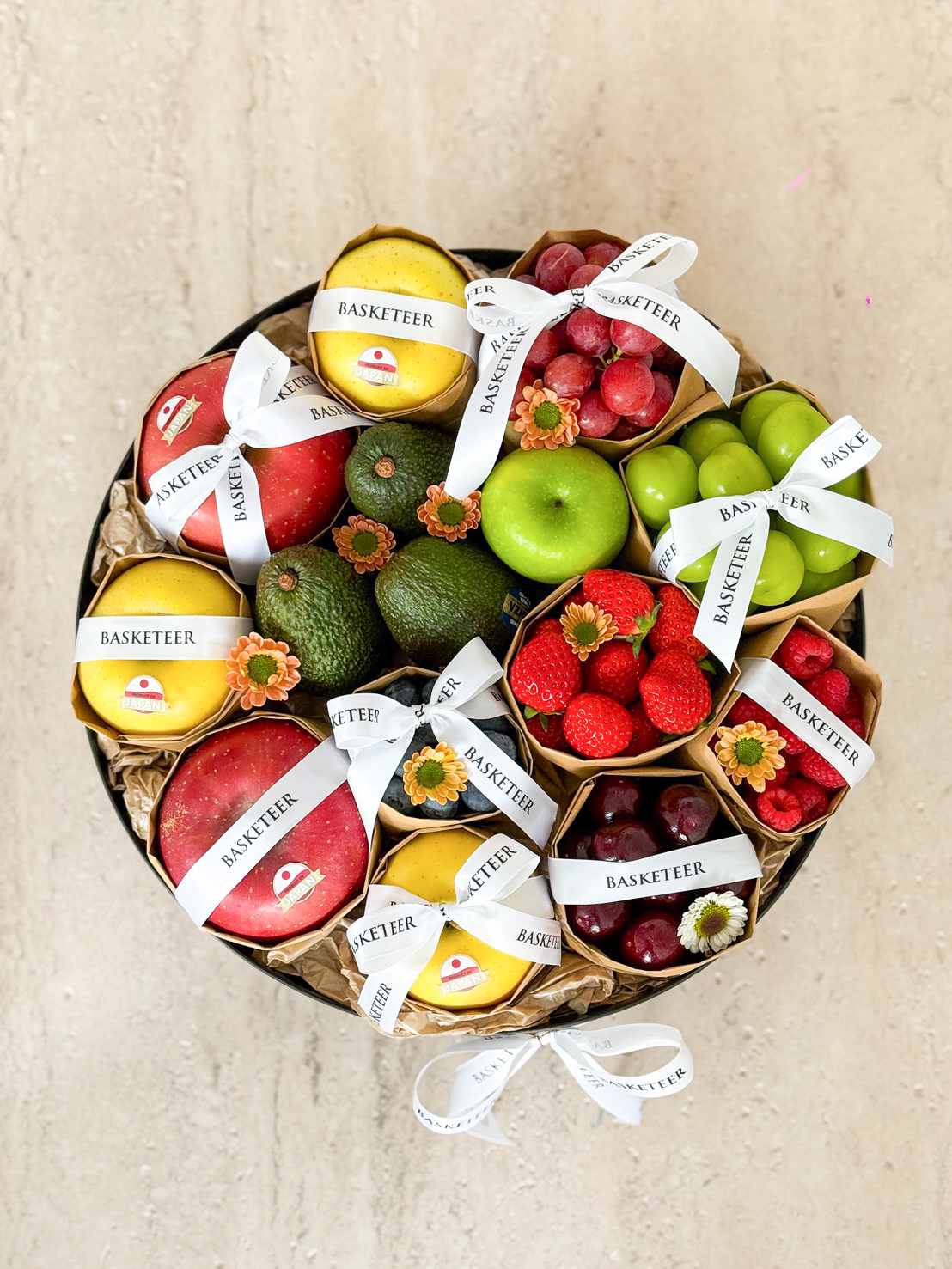 A Mixed Fruit Round Gift Box filled with assorted fruits—apples, avocados, grapes, strawberries, lemons, and raspberries—beautifully arranged in a round basket with white 