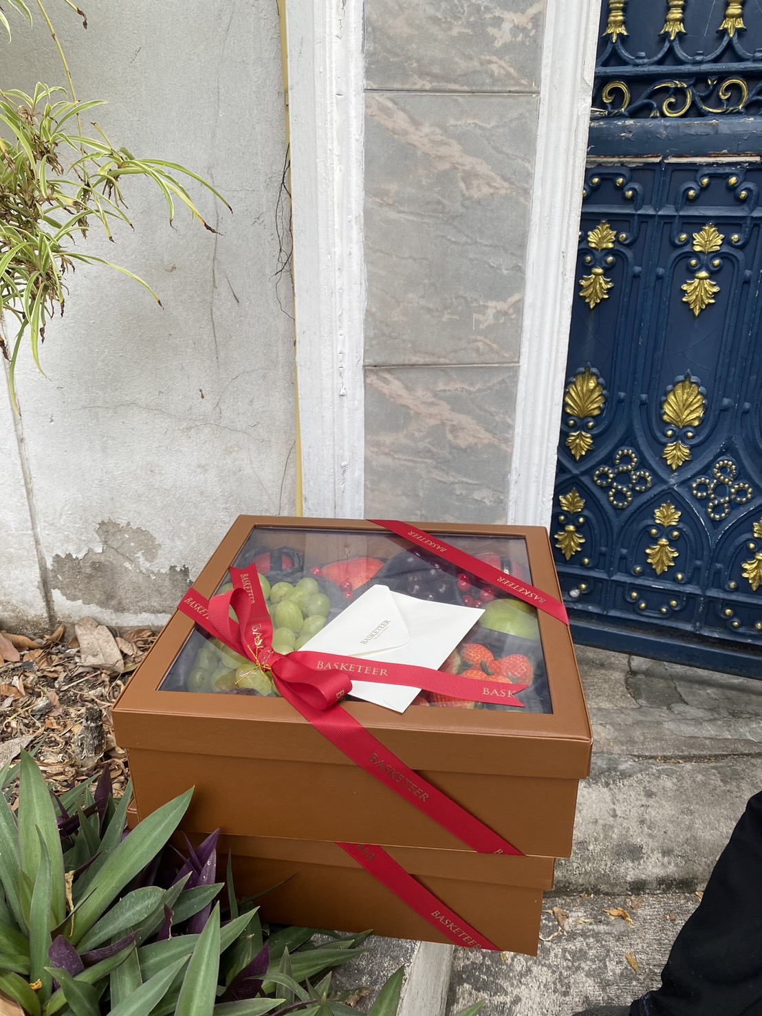 A neatly stacked pair of brown gift boxes with transparent lids, filled with fresh fruits like grapes and strawberries, wrapped with elegant red ribbons and accompanied by a card, placed near a decorative blue gate in an outdoor setting.