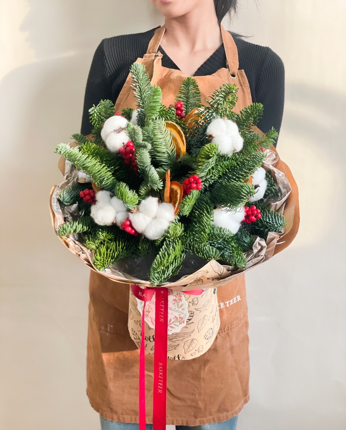 Festive wicker basket decorated with fresh white roses, cotton blooms, green grapes, and a green ribbon adorned with a Christmas tree tag.