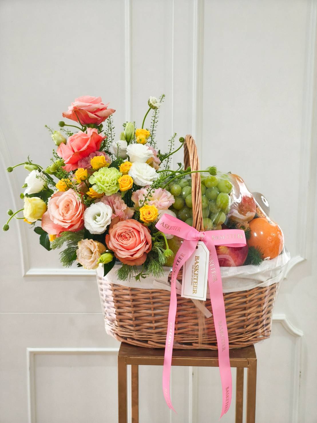 Luxury fruit and flower basket with green grapes, apples, and assorted fruits, decorated with peach and pink roses, white lisianthus, and yellow spray roses, tied with a pink satin ribbon.