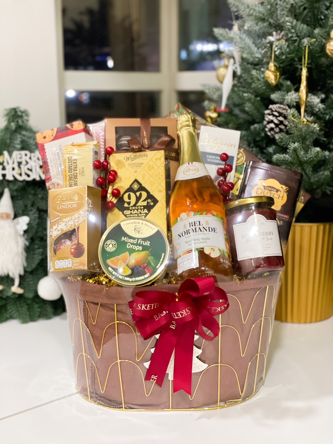 A luxurious Christmas gift basket by Basketeer, filled with premium treats such as chocolates, mixed fruit drops, sparkling apple cider, and gourmet snacks, beautifully arranged in a gold-accented basket with a festive red ribbon, set in a cozy holiday scene with a Christmas tree in the background.