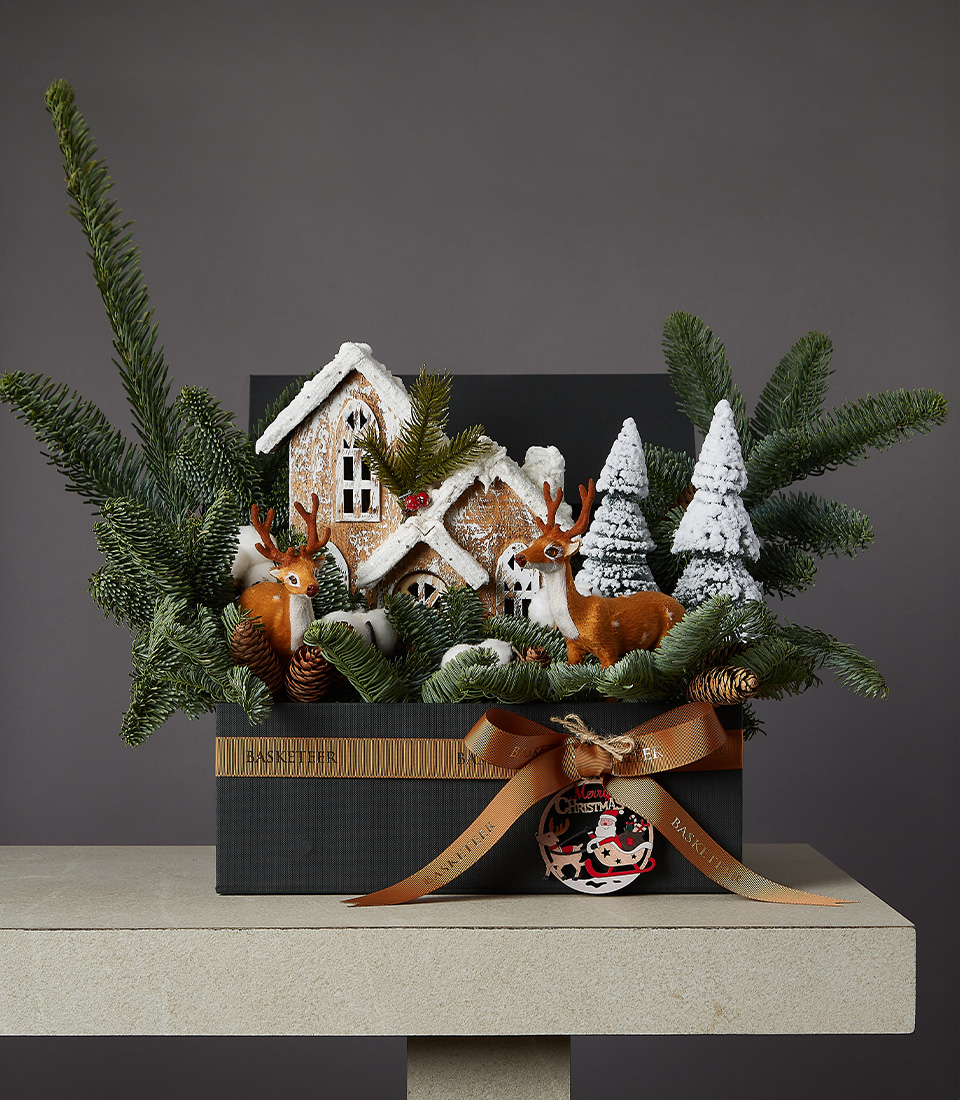 Festive Christmas arrangement with reindeer, cottage, evergreen branches, and snowy decorations in a gift box.