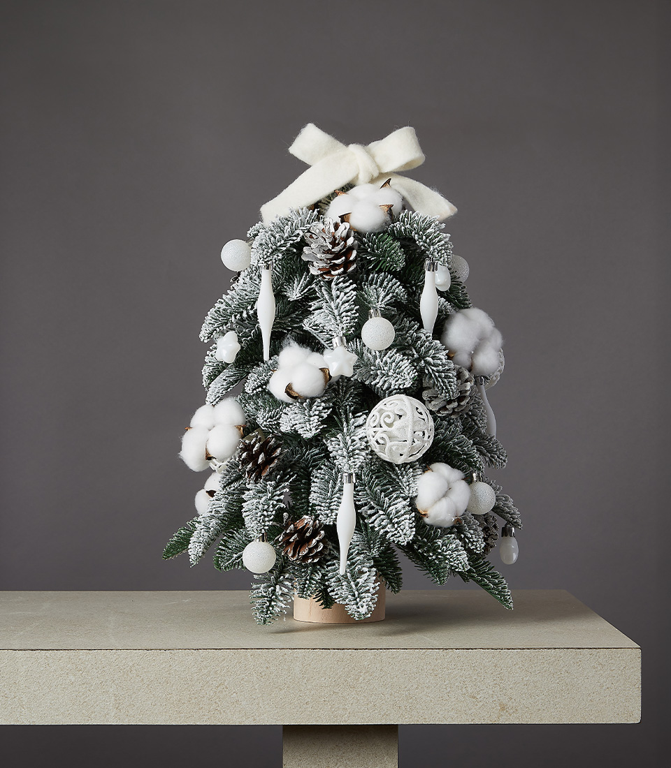 Snowy Christmas tree arrangement with frosted evergreen branches, cotton accents, pinecones, and white decorations topped with a felt bow.