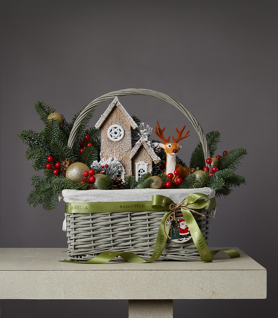 Christmas basket with reindeer figurine, frosted pine branches, red berries, and rustic wooden houses.