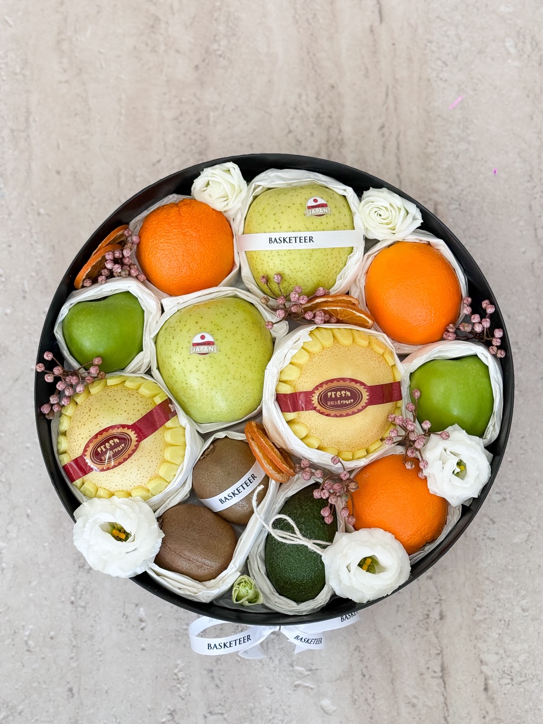 An Elegant Citrus & Pear Gift Hamper with Kiwi, this round black hamper brims with pears, oranges, apples, avocado, and kiwi, adorned with white flowers, pink berries, and a 