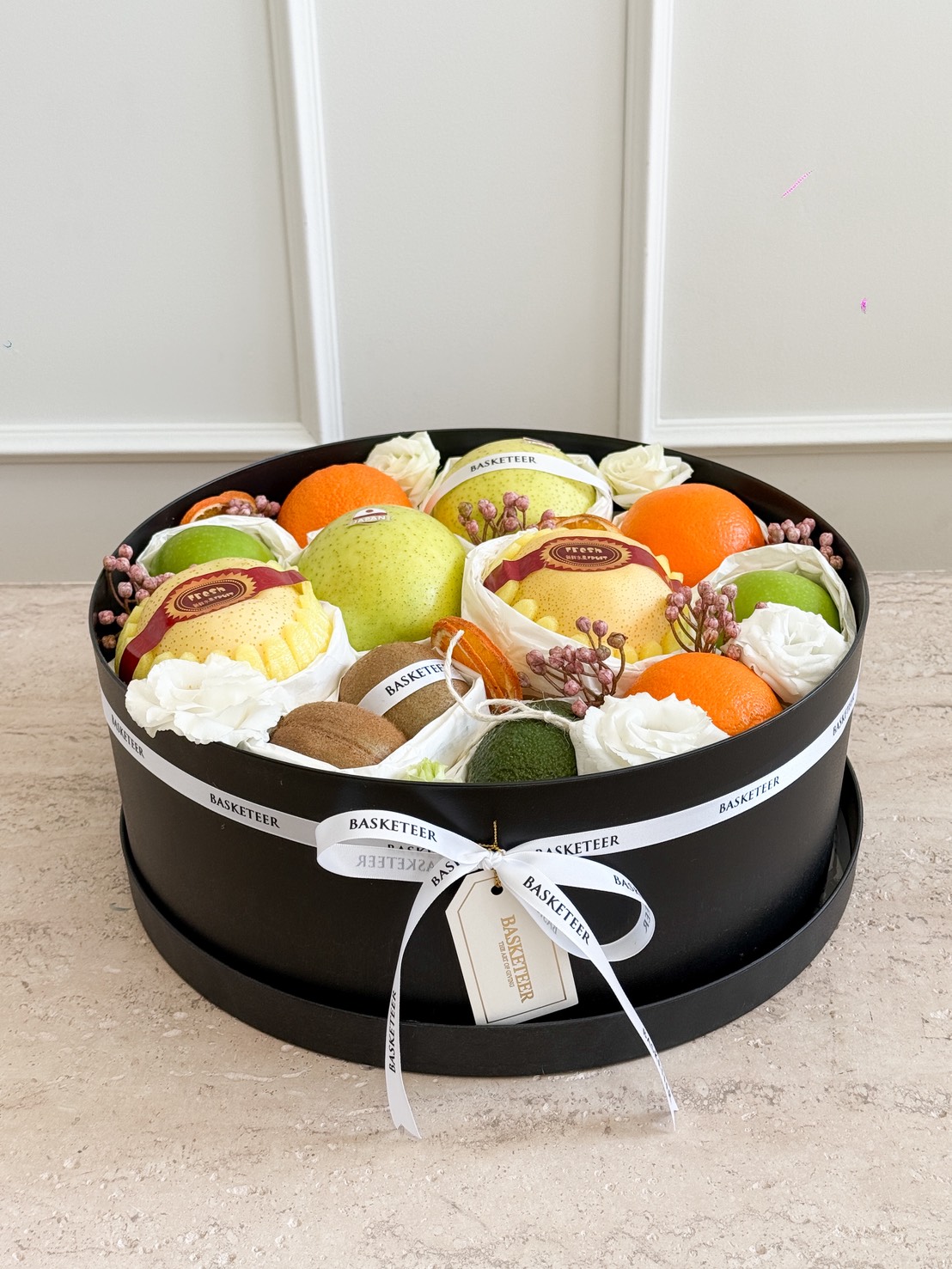 An Elegant Citrus & Pear Gift Hamper with Kiwi, this round black box is filled with fresh apples, oranges, kiwi, and pears, beautifully decorated with white artificial flowers and a “Basketeer” ribbon.