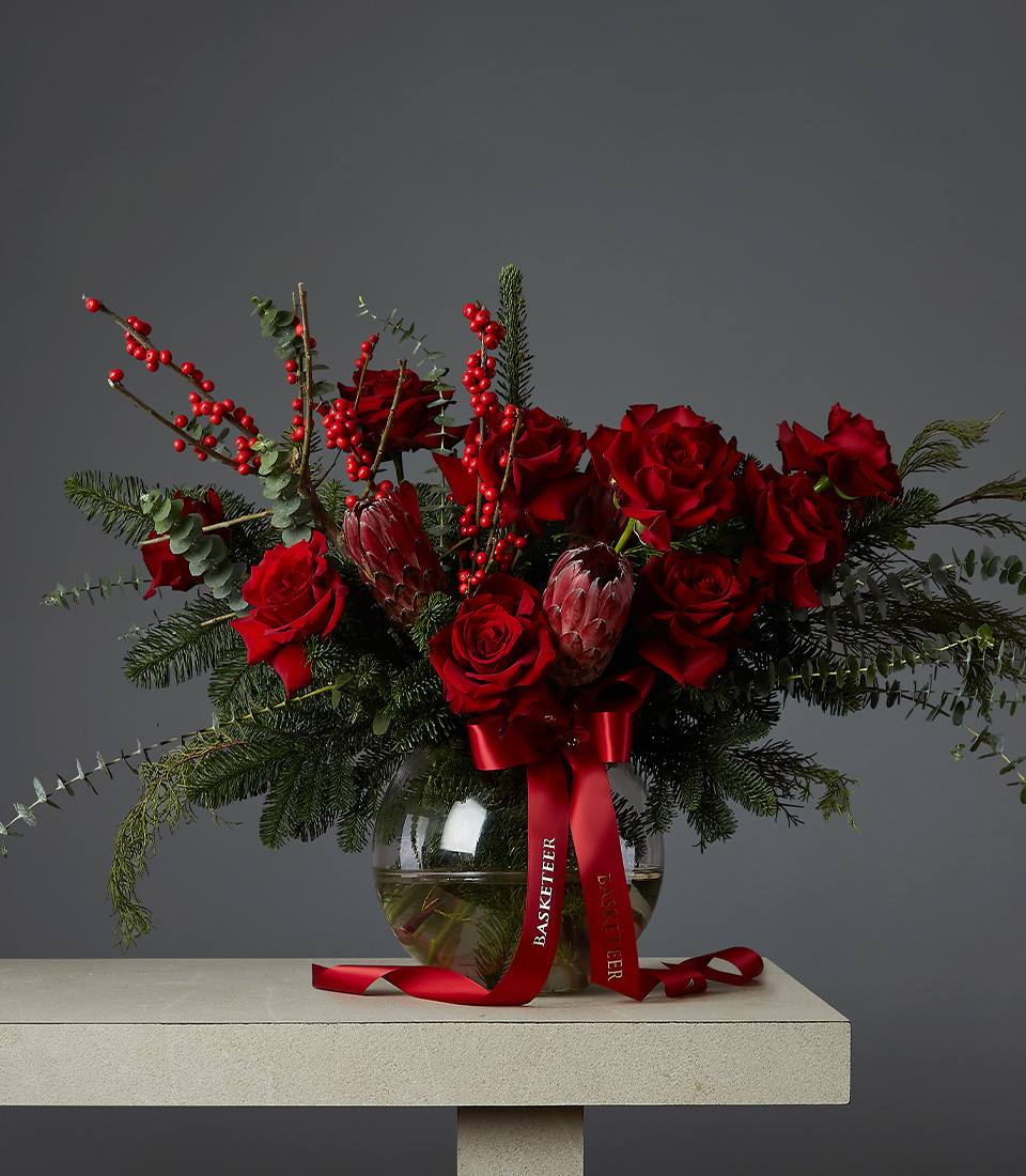 A bouquet of red roses, berries, and protea flowers arranged with greenery in a clear glass vase. The vase sits on a light-colored table against a dark gray background. A red ribbon with text is tied around the vase.
