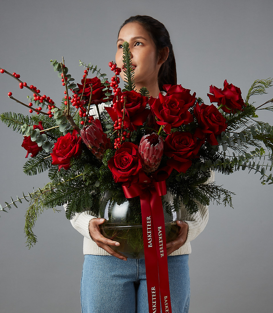A woman in a white sweater and blue jeans holds a large bouquet of red roses, greenery, and red berries in a clear round vase. A red ribbon with 