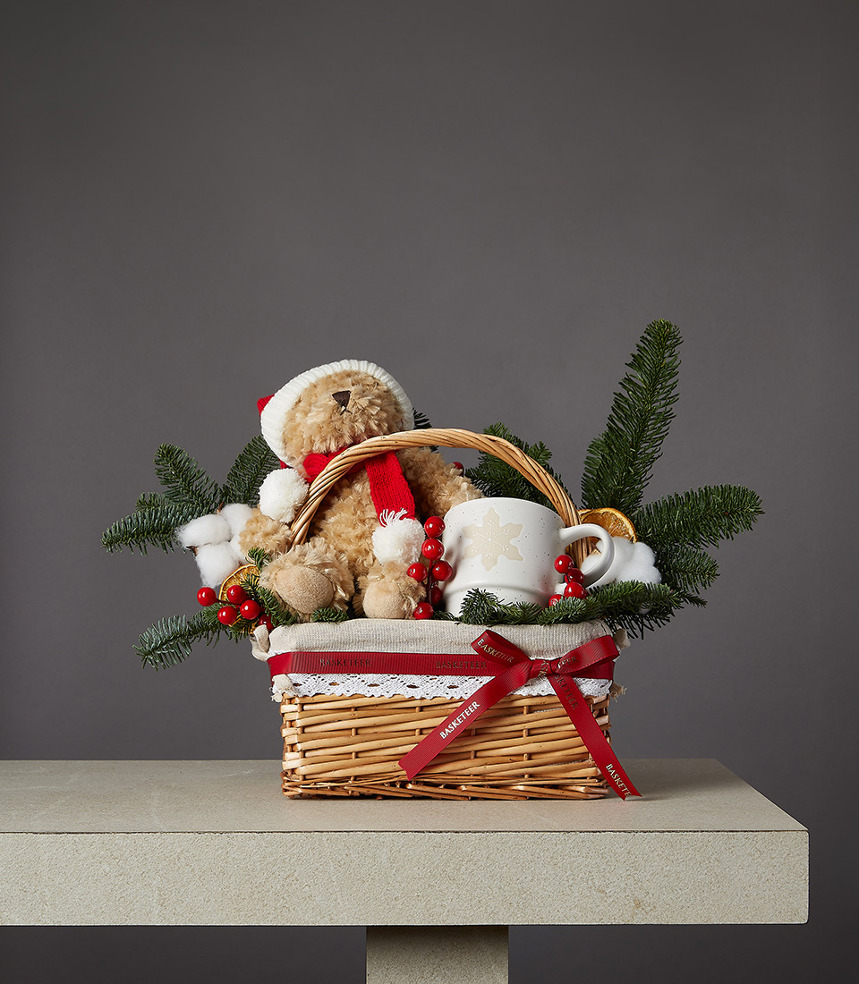 Wicker Christmas basket with a teddy bear in festive attire, holiday greenery, a ceramic mug, and red decorative accents.