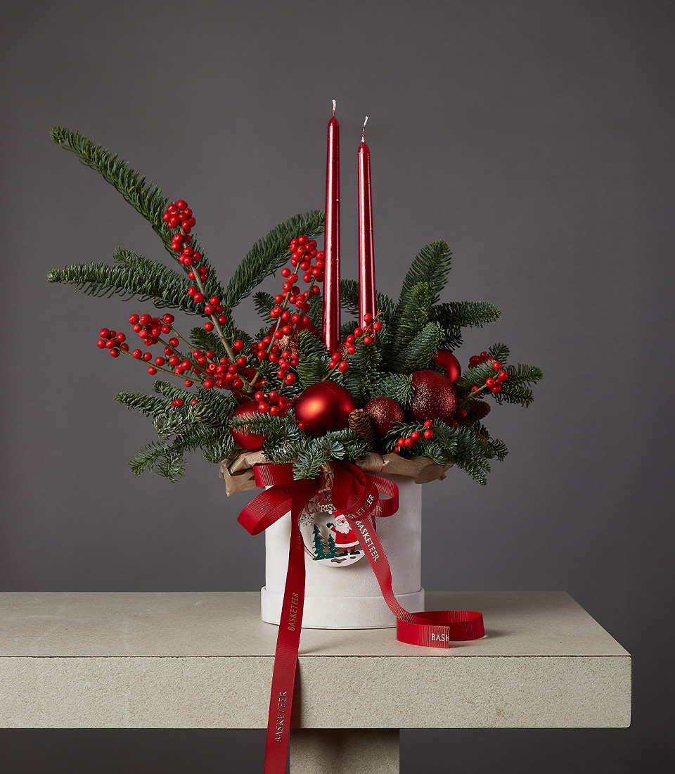 A luxurious floral arrangement featuring deep red roses, proteas, festive berries, and greenery in a clear glass vase with a red ribbon.