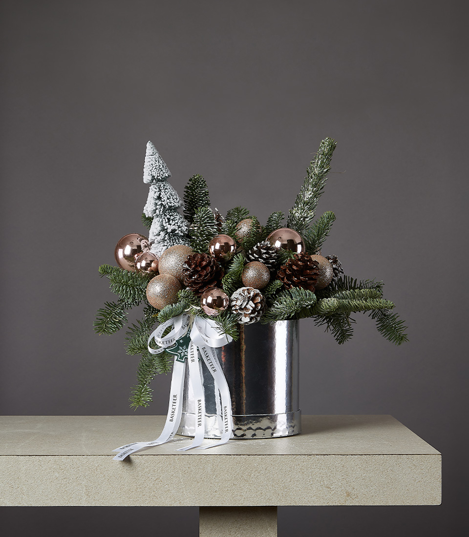 Christmas arrangement with frosted pine branches, bronze ornaments, pinecones, and a snowy tree in a silver vase.