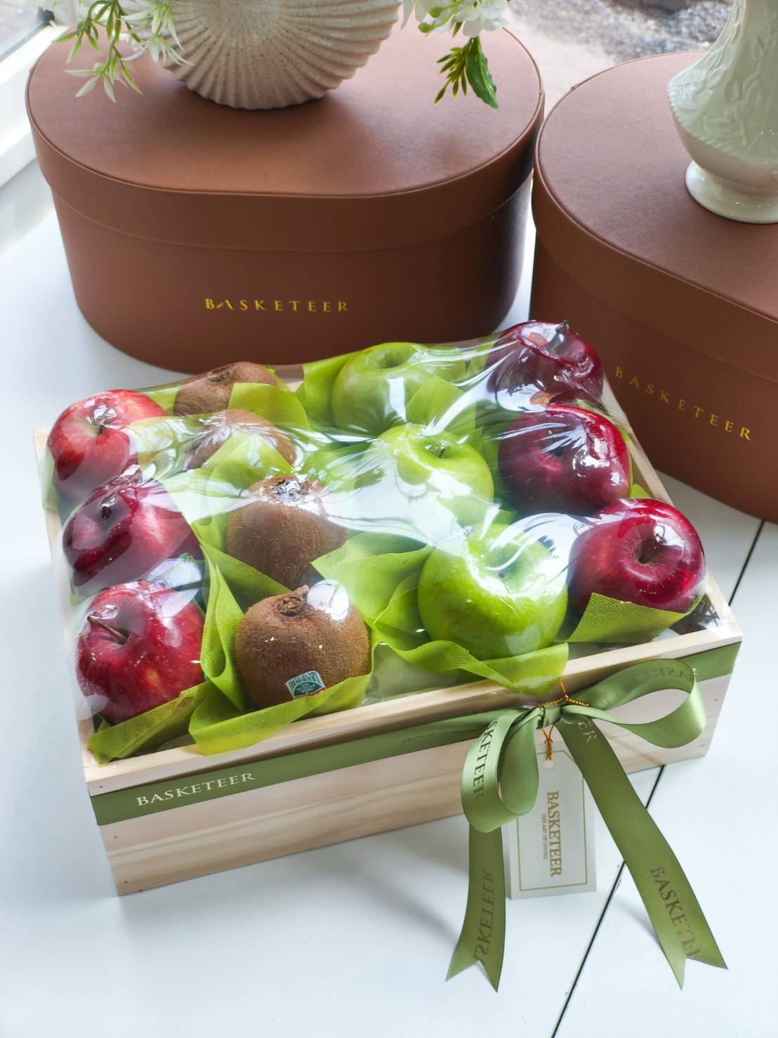 Premium fruit gift box with fresh red apples, green apples, and kiwis arranged in a wooden box with green ribbon decoration.