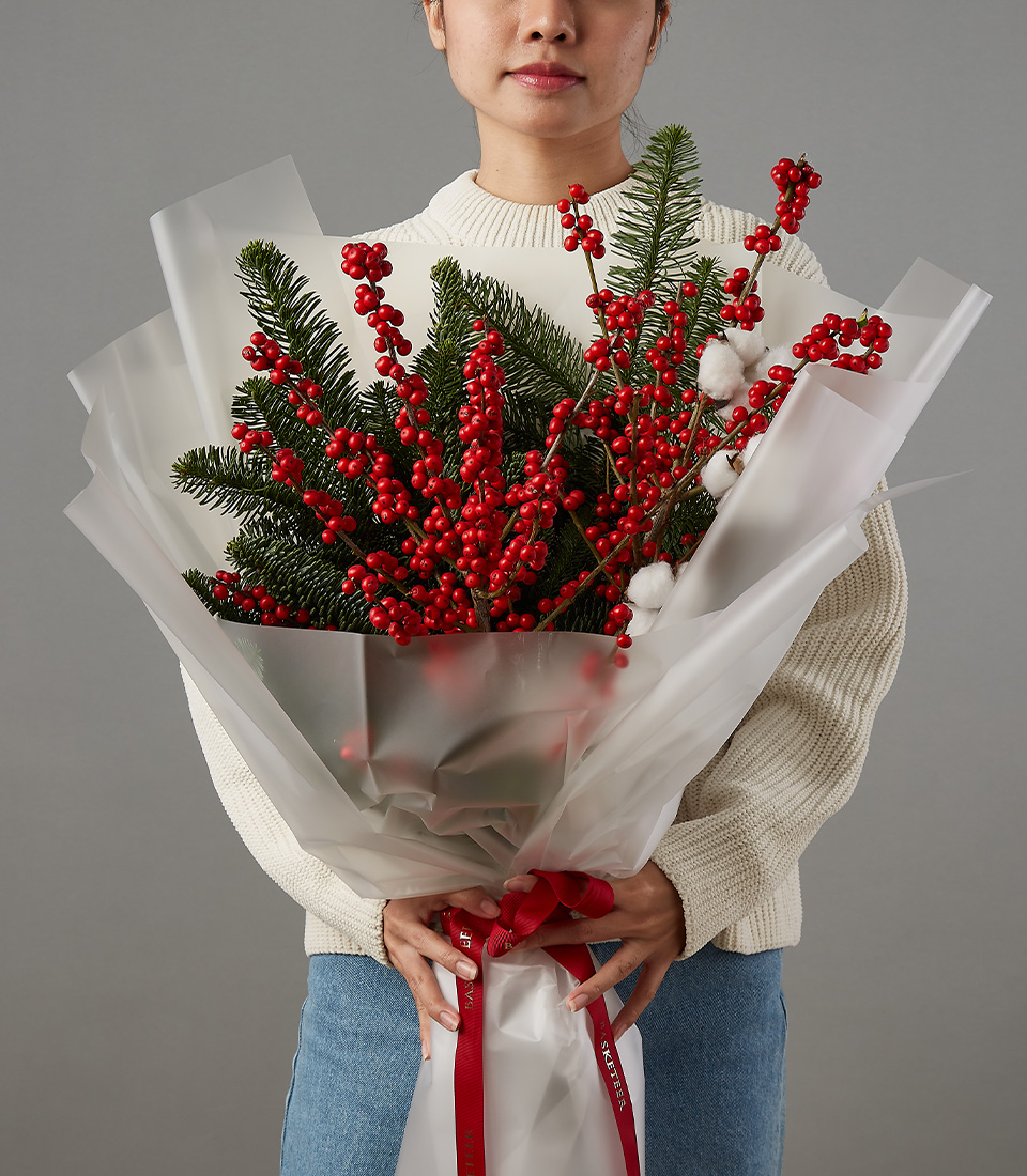 Christmas bouquet featuring vibrant red berries, greenery, and cotton accents, wrapped in translucent paper, held by a person in a cream sweater.