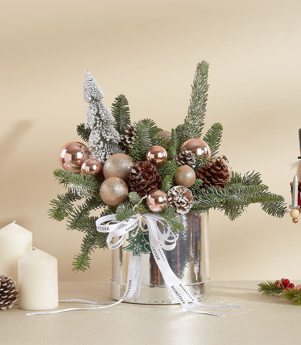 Holiday arrangement with pine branches, rose gold ornaments, pinecones, and a snow-dusted tree in a silver container.