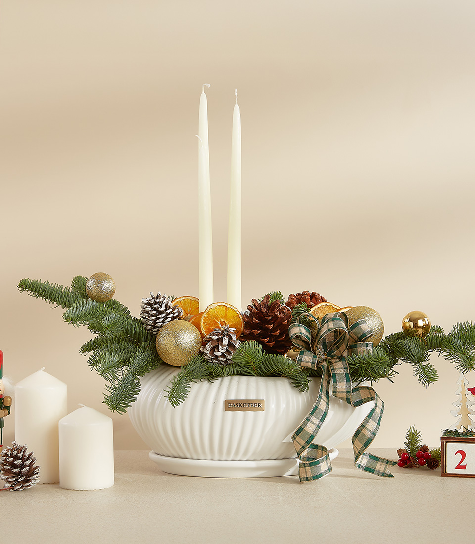 Holiday centerpiece with pine branches, pinecones, gold ornaments, and candles in a white ceramic vase.