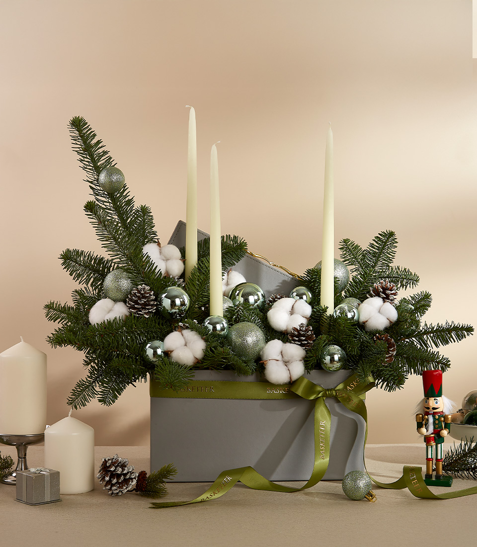 Holiday centerpiece with pine branches, cotton accents, silver baubles, and three ivory candles in a grey box.