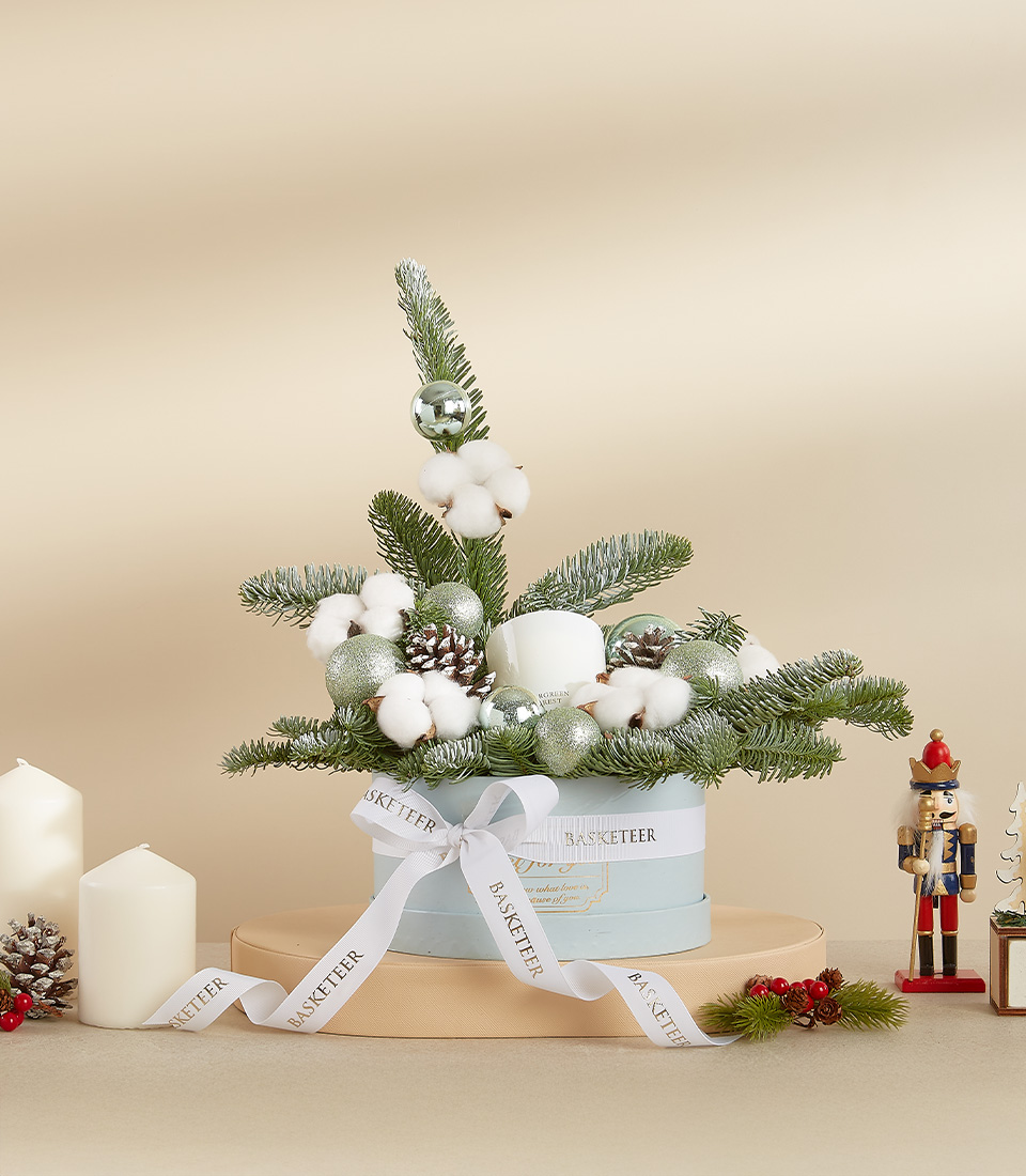 Elegant holiday arrangement with frosted pine branches, cotton accents, silver baubles, and pinecones in a pastel blue hatbox.