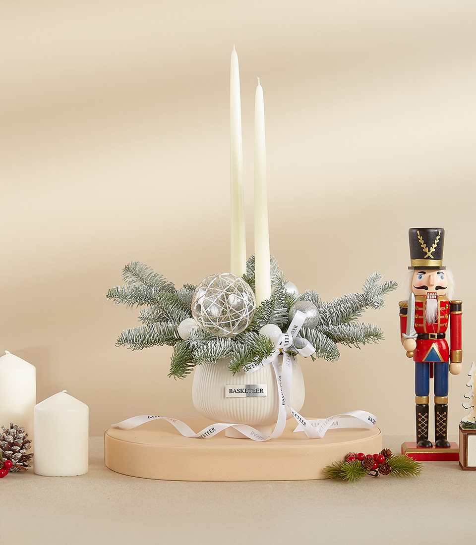 Holiday candle centerpiece with frosted pine branches, silver baubles, and two white taper candles in a ceramic base.