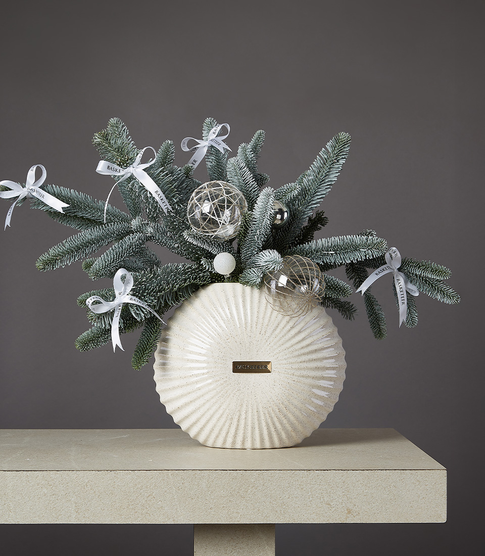 Christmas arrangement with frosted pine branches, silver ornaments, and a white ceramic vase on a beige table.