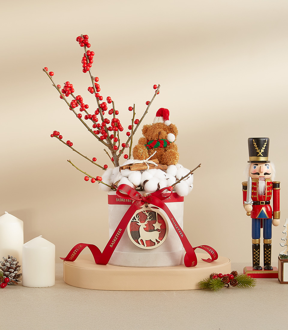 Christmas arrangement featuring a teddy bear with red berry branches, cotton accents, and a holiday ribbon in a decorative box.