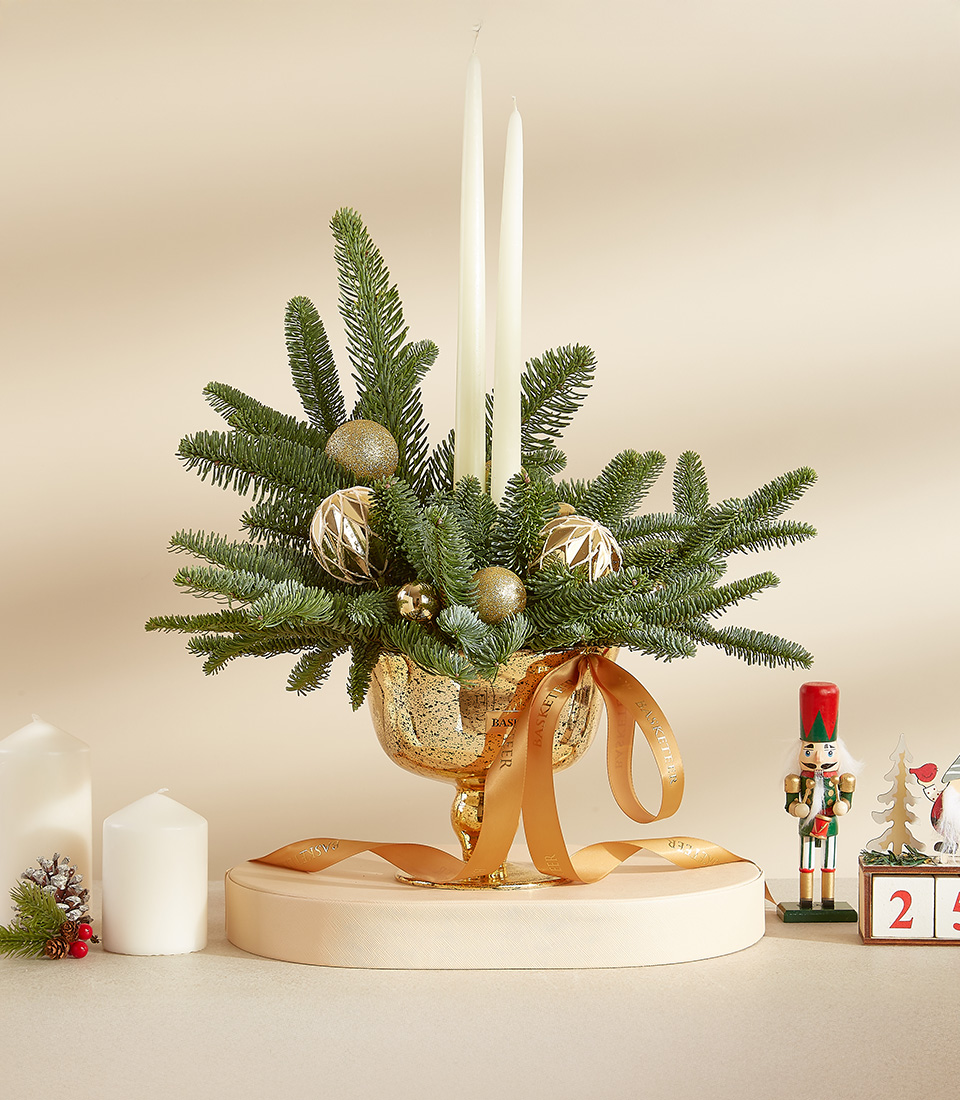 Christmas candle arrangement with white candles, greenery, and gold ornaments in a golden vase, accented with decorative ribbons.