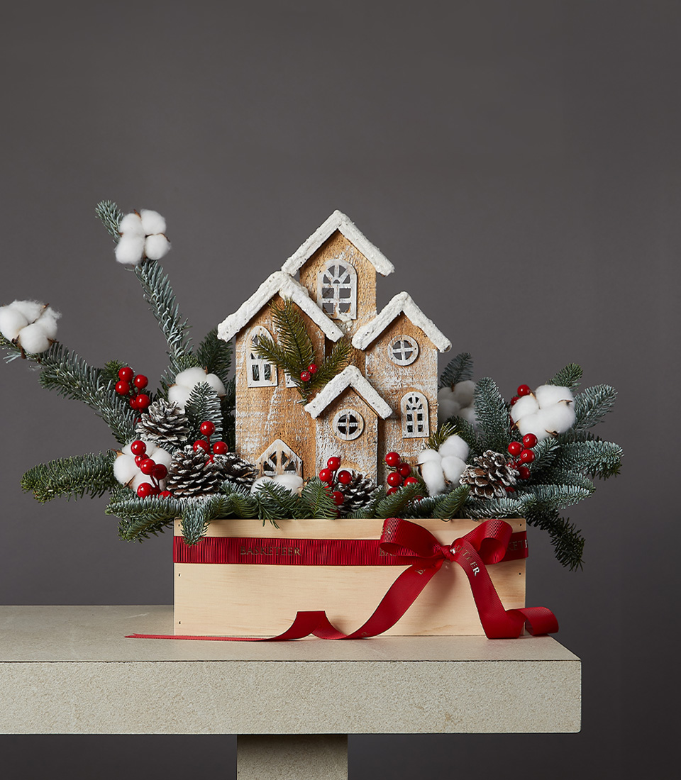 Snowy Village Christmas centerpiece featuring a charming wooden village with snowy roofs, surrounded by pine branches, pine cones, red berries, and fluffy cotton flowers. All beautifully set in a wooden box with a red ribbon on a light-colored table.