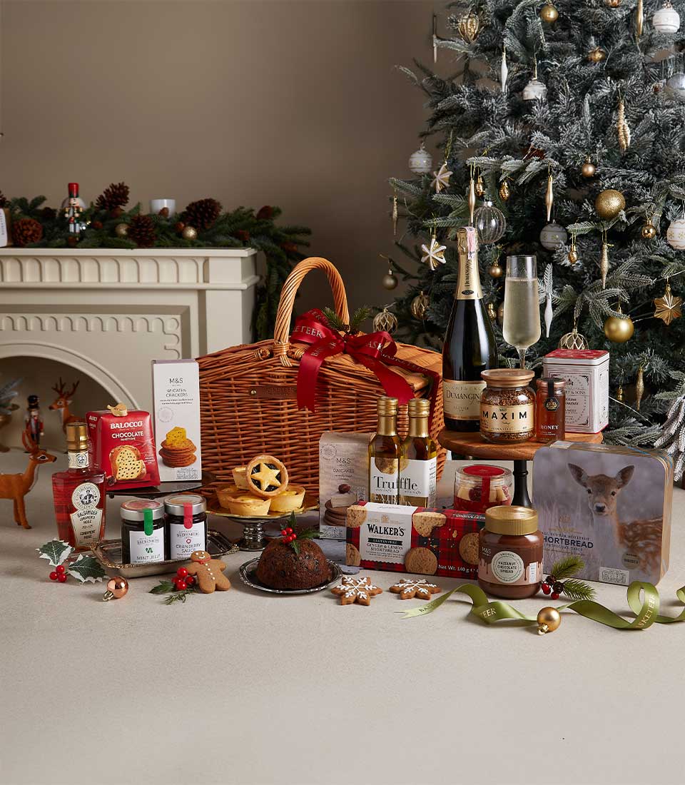 Wicker Christmas hamper filled with gourmet holiday treats, cookies, and preserves, placed near a Christmas tree and festive decorations.