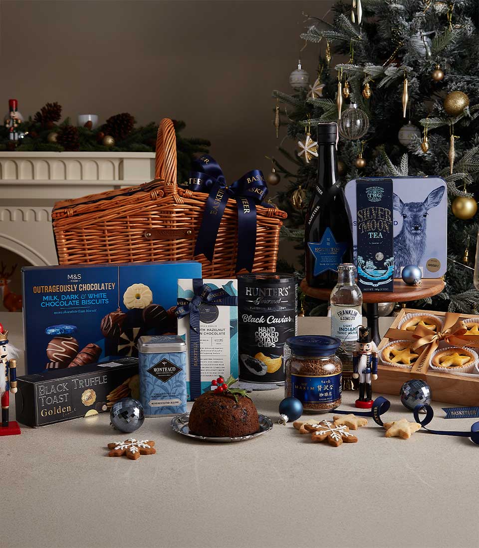 Midnight blue-themed Christmas hamper with fine teas, chocolates, truffle snacks, and festive treats near a decorated Christmas tree.