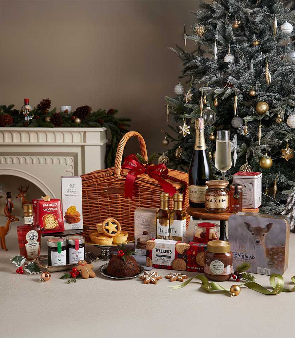 Wicker Christmas hamper filled with gourmet holiday treats, cookies, and preserves, placed near a Christmas tree and festive decorations.