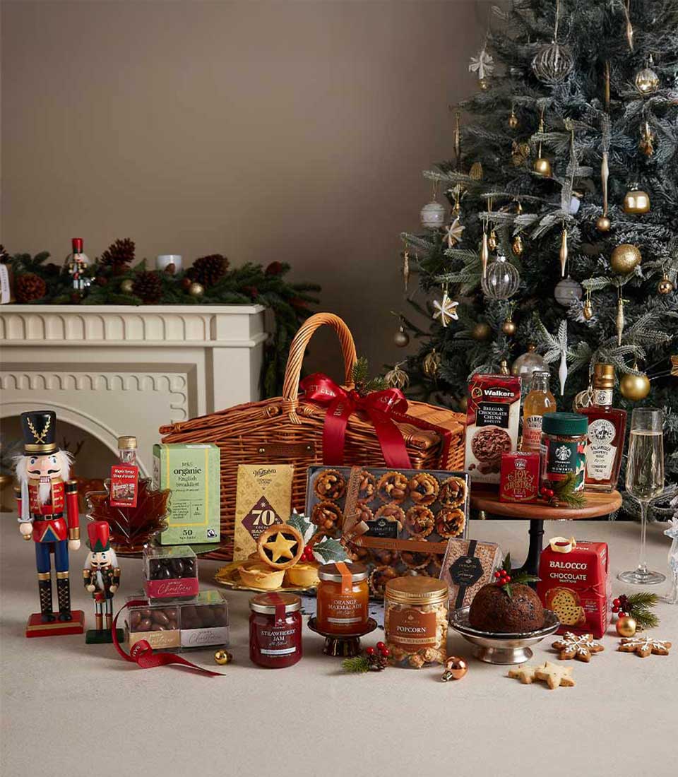 Grand Christmas hamper with an assortment of chocolates, cookies, preserves, and holiday treats, set near a Christmas tree and fireplace.
