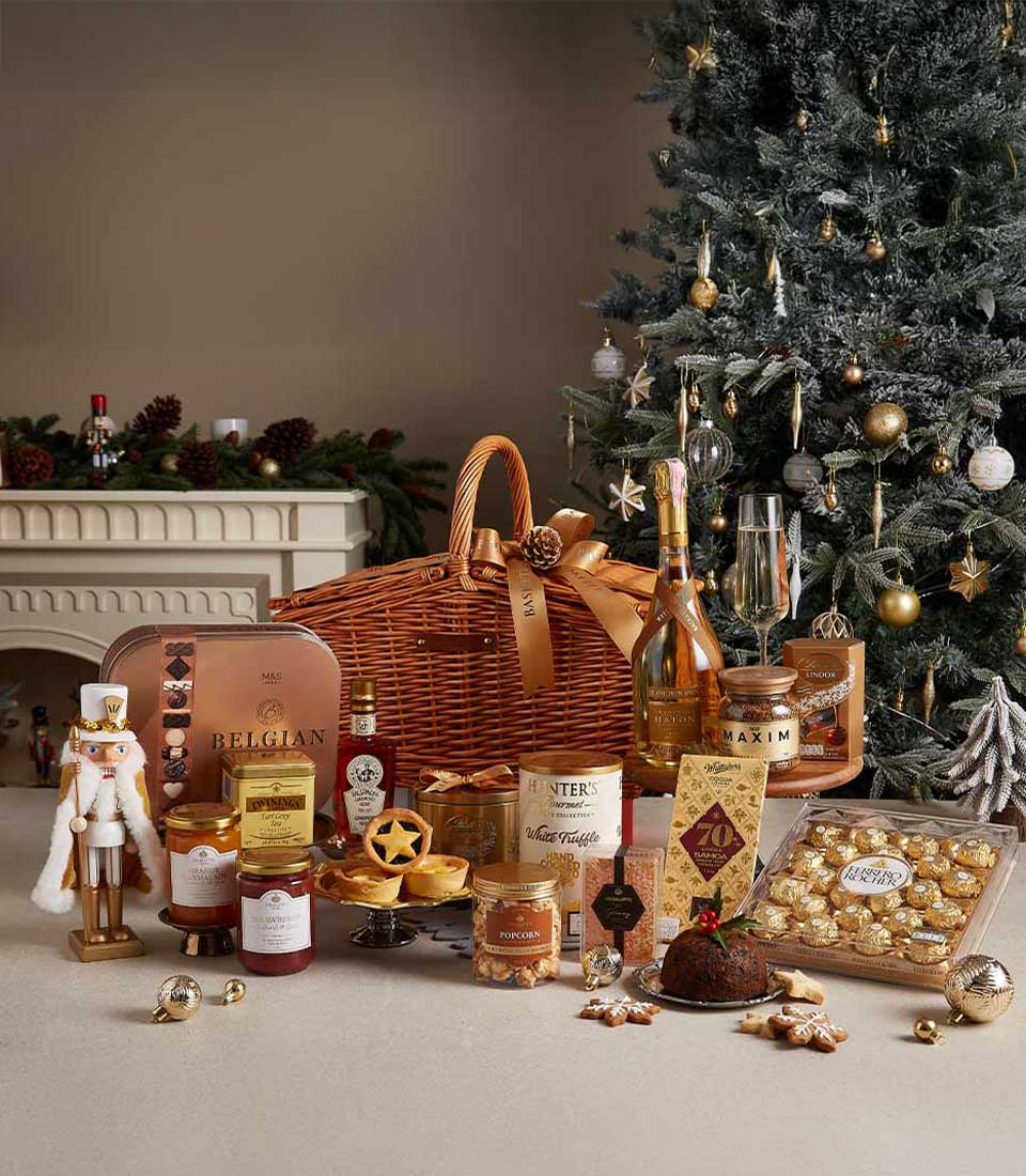 Premium wicker Christmas hamper filled with Belgian chocolates, gourmet preserves, popcorn, and holiday treats, displayed near a decorated Christmas tree.