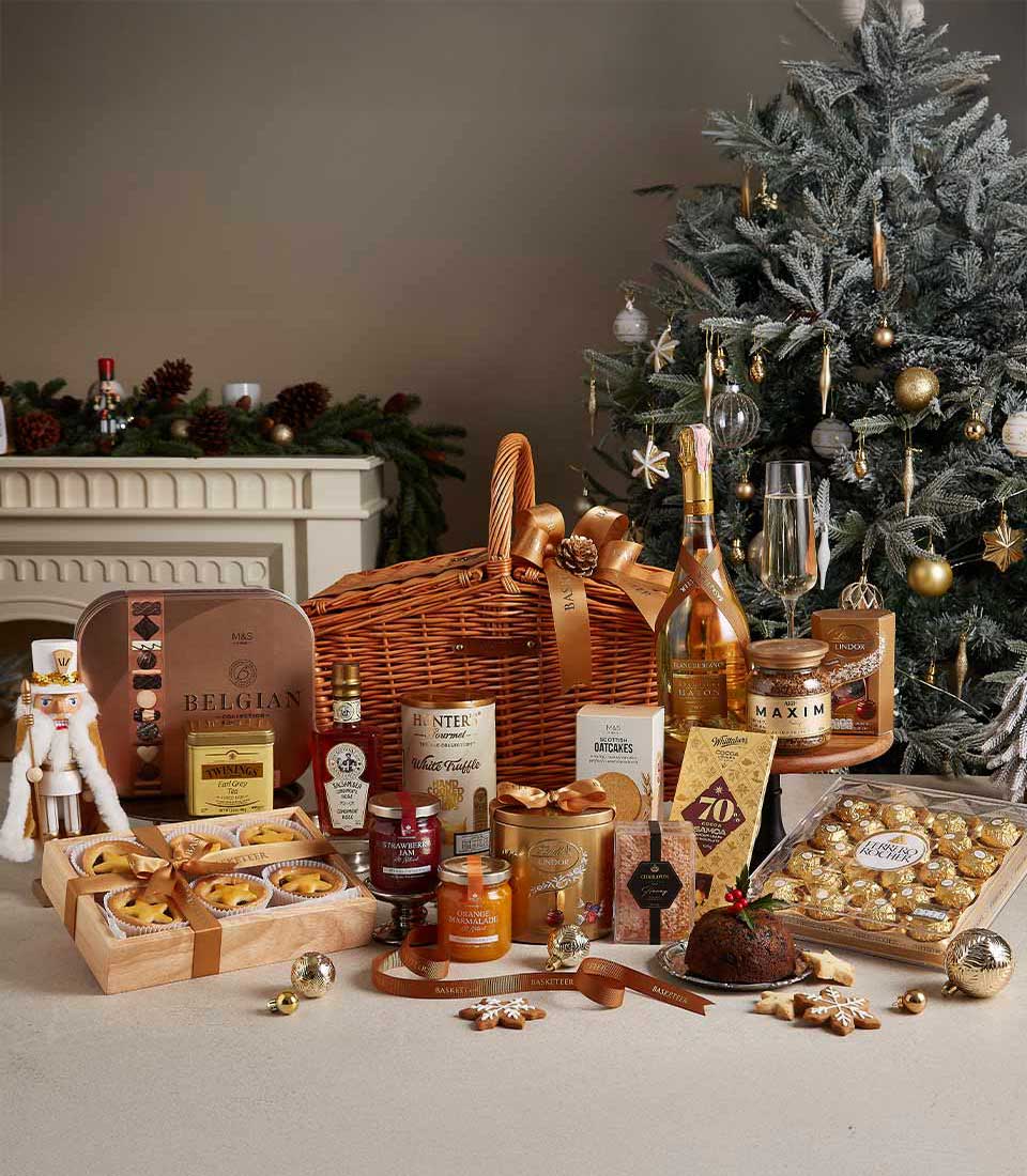 Deluxe Christmas hamper with Belgian chocolates, festive pastries, preserves, and holiday treats, arranged near a decorated Christmas tree.