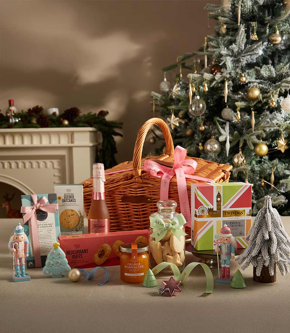 A wicker gift basket adorned with a pink ribbon, filled with Twinings tea, Freixenet sparkling rosé, M&S cookies, redcurrant puffs, orange marmalade, and festive decorations, set by a Christmas tree.
