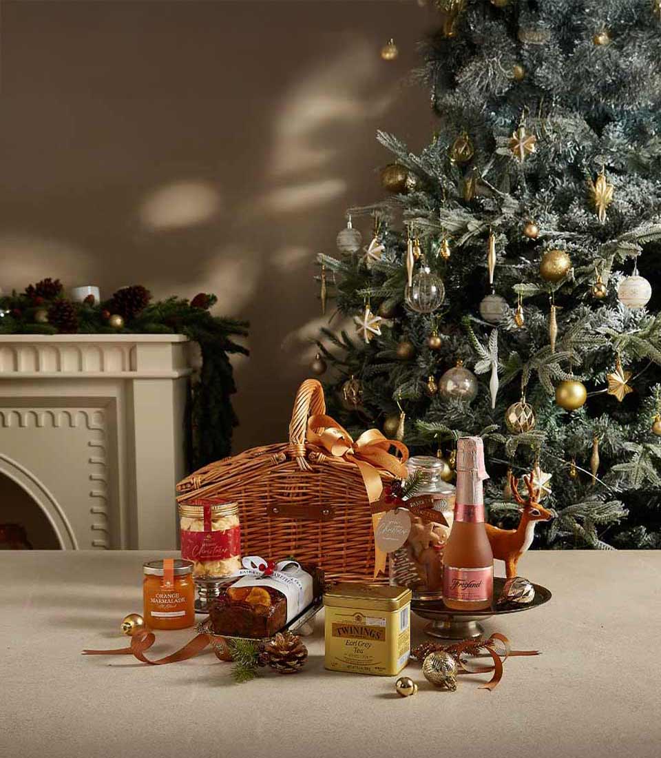 Elegant Christmas hamper with Earl Grey tea, orange marmalade, fruit cake, and Freixenet Rosé, displayed near a beautifully decorated Christmas tree.