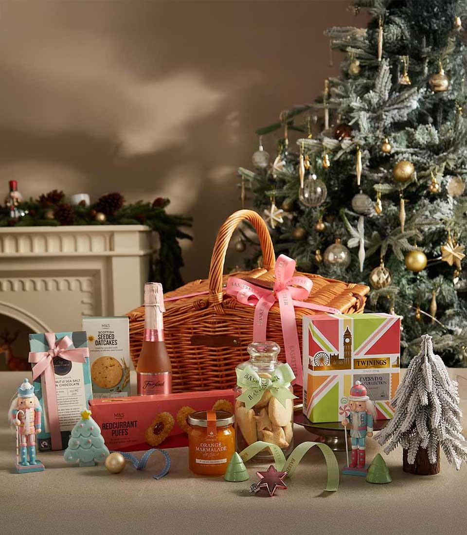 A wicker gift basket adorned with a pink ribbon, filled with Twinings tea, Freixenet sparkling rosé, M&S cookies, redcurrant puffs, orange marmalade, and festive decorations, set by a Christmas tree.