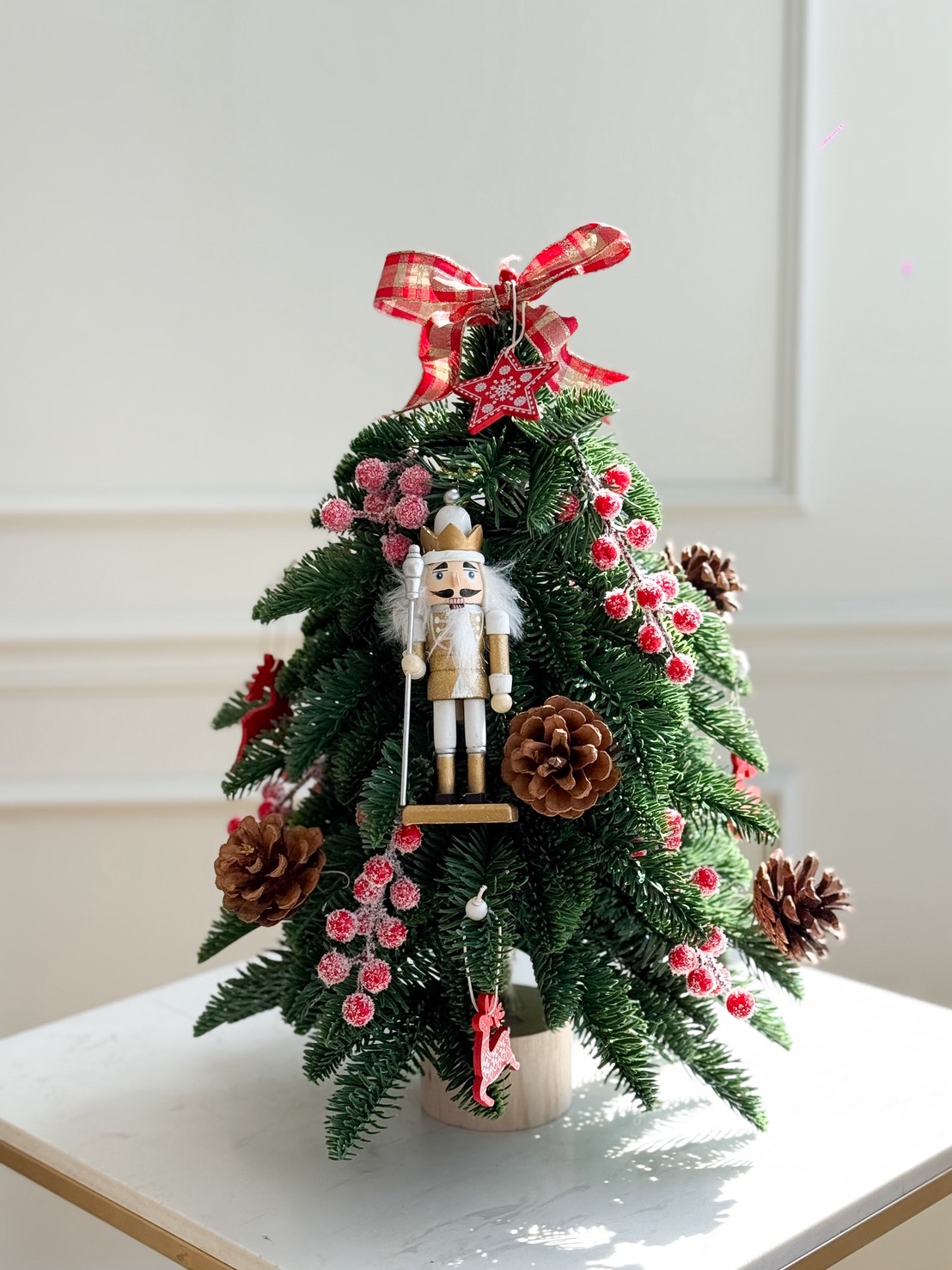 A Nutcracker Mini Christmas Tree adorned with pinecones, red berries, a checked ribbon on top, and a wooden nutcracker ornament hanging in the centre sits on a white table in a bright room.