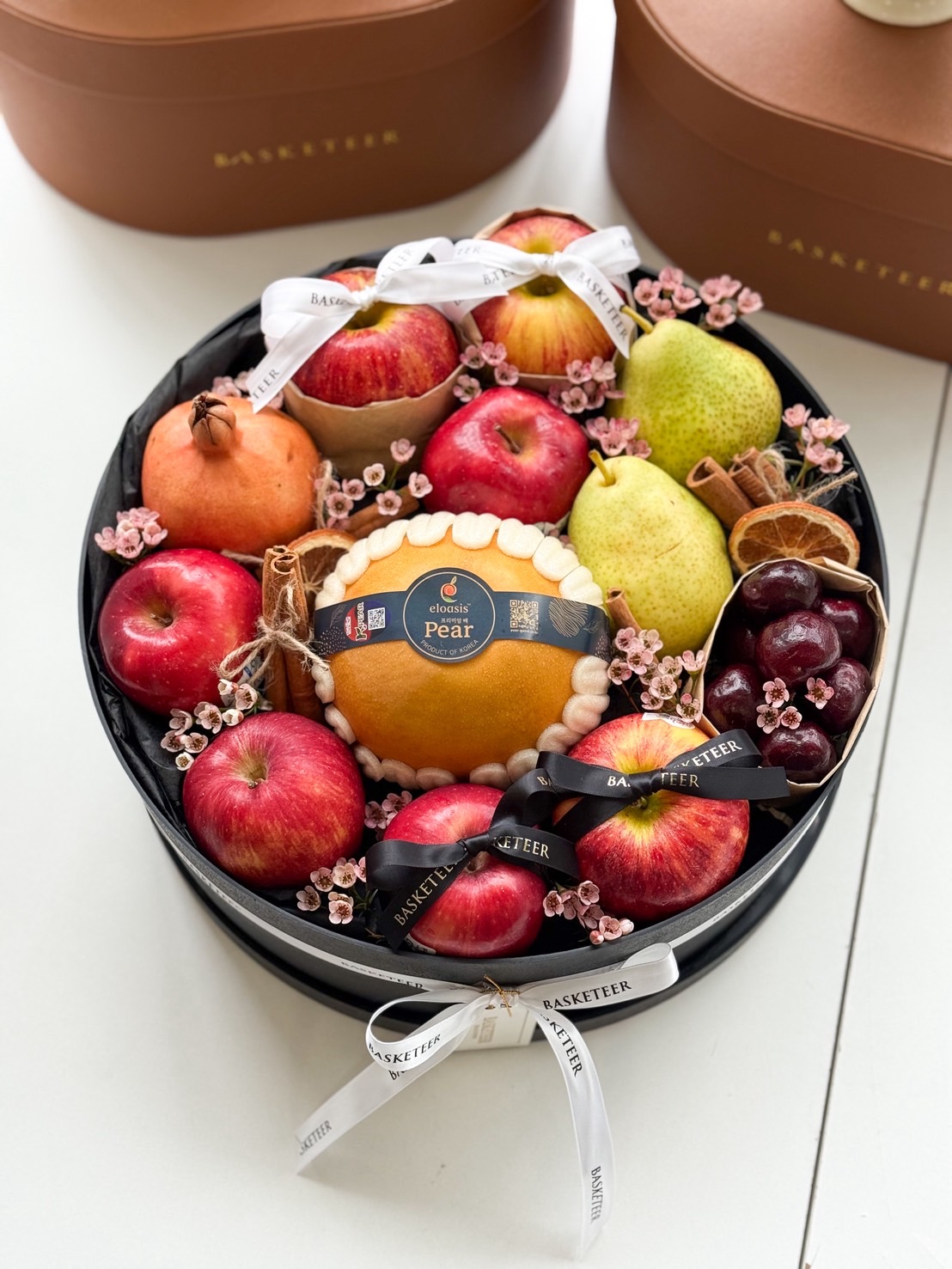 Luxury black round box filled with premium apples, pears, pomegranates, grapes, and Korean pears, accented with pink flowers.