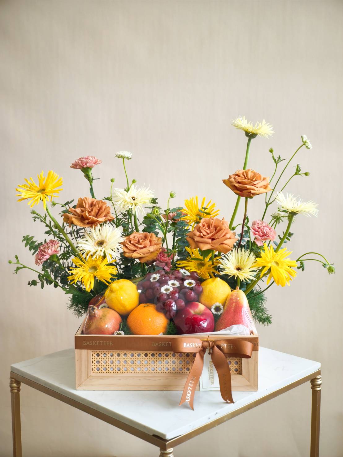 Wooden basket with fresh fruits including grapes, oranges, pears, and lemons, decorated with yellow, white, and peach flowers.