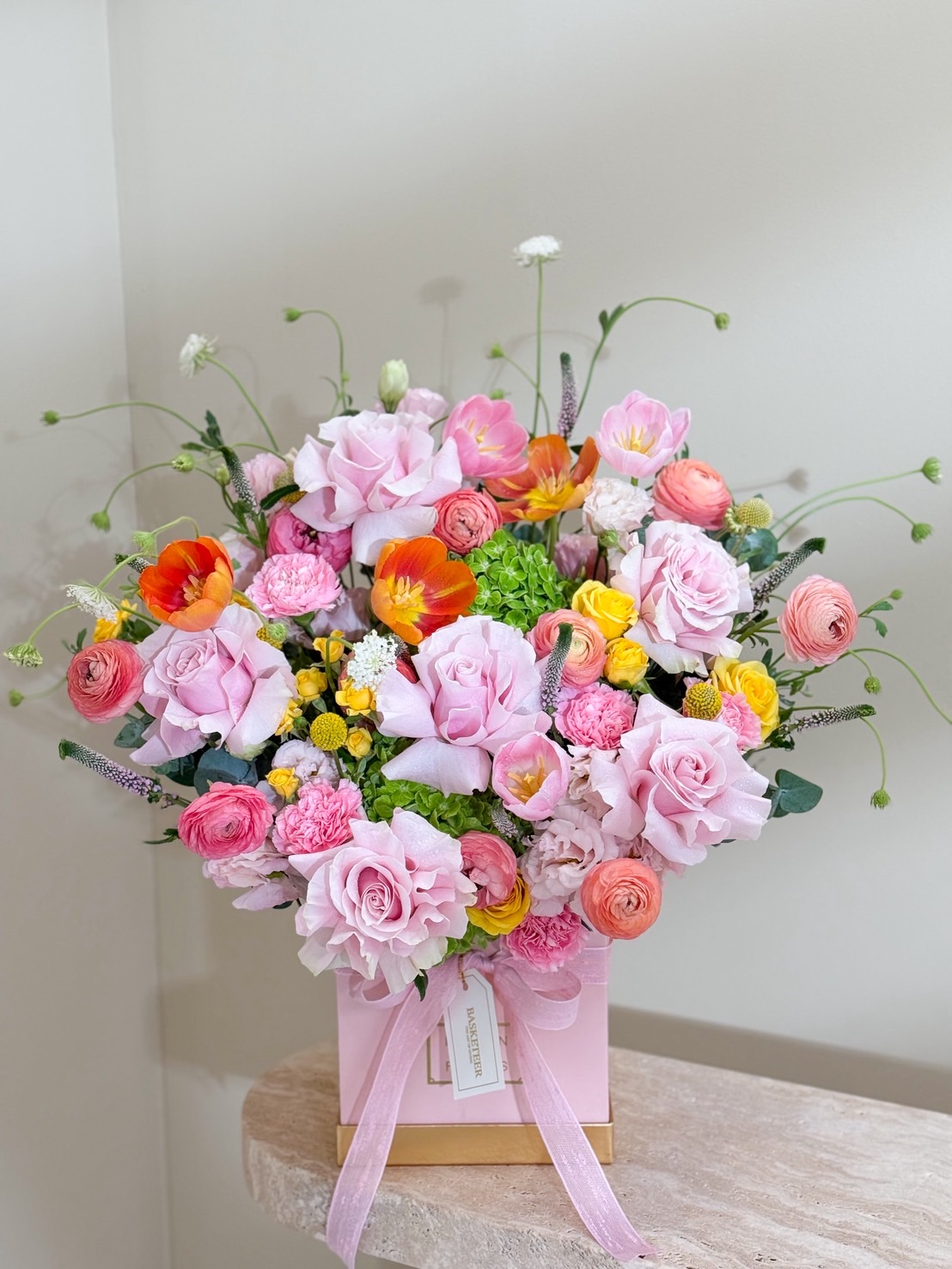 Vibrant pink flower arrangement in a pastel pink box with roses, ranunculus, and mixed blooms tied with ribbon.