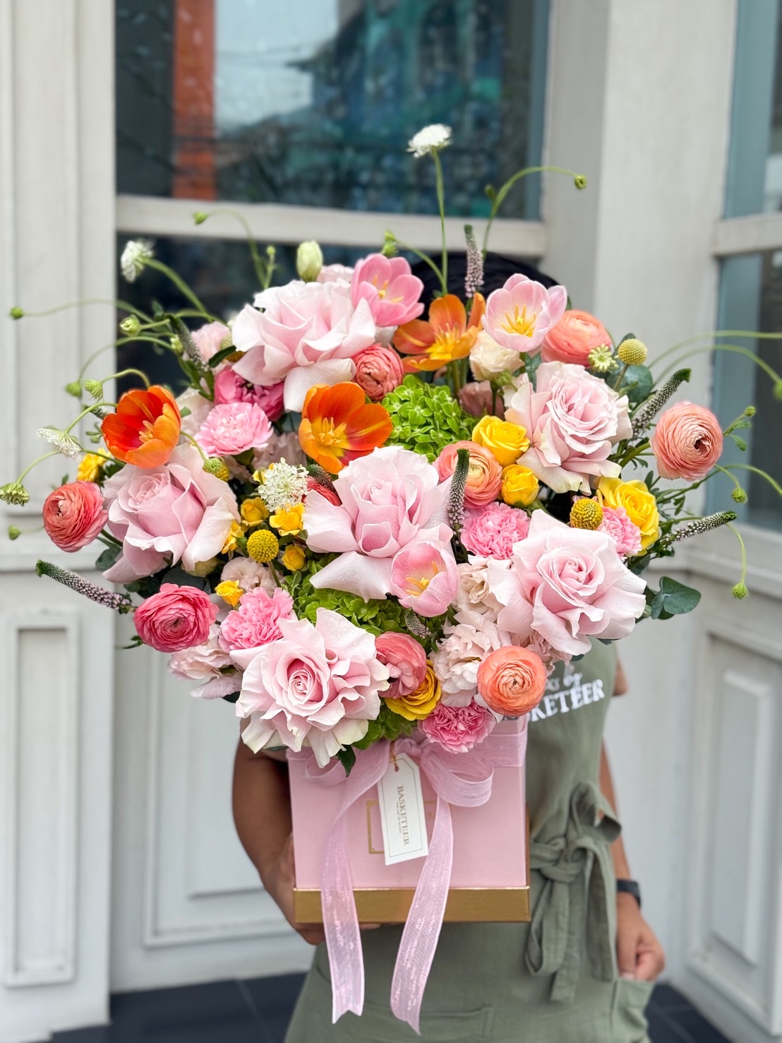 Vibrant pink flower arrangement in pastel pink gift box with roses, ranunculus, orange and yellow mixed blooms held outdoors.